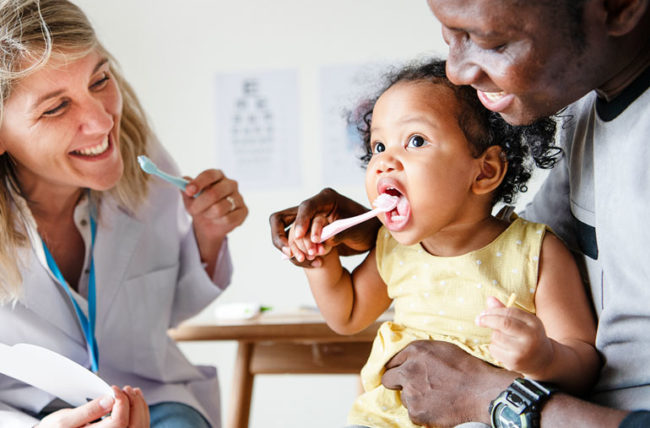 A baby brushing her teeth with her dentist and a parent nearby.